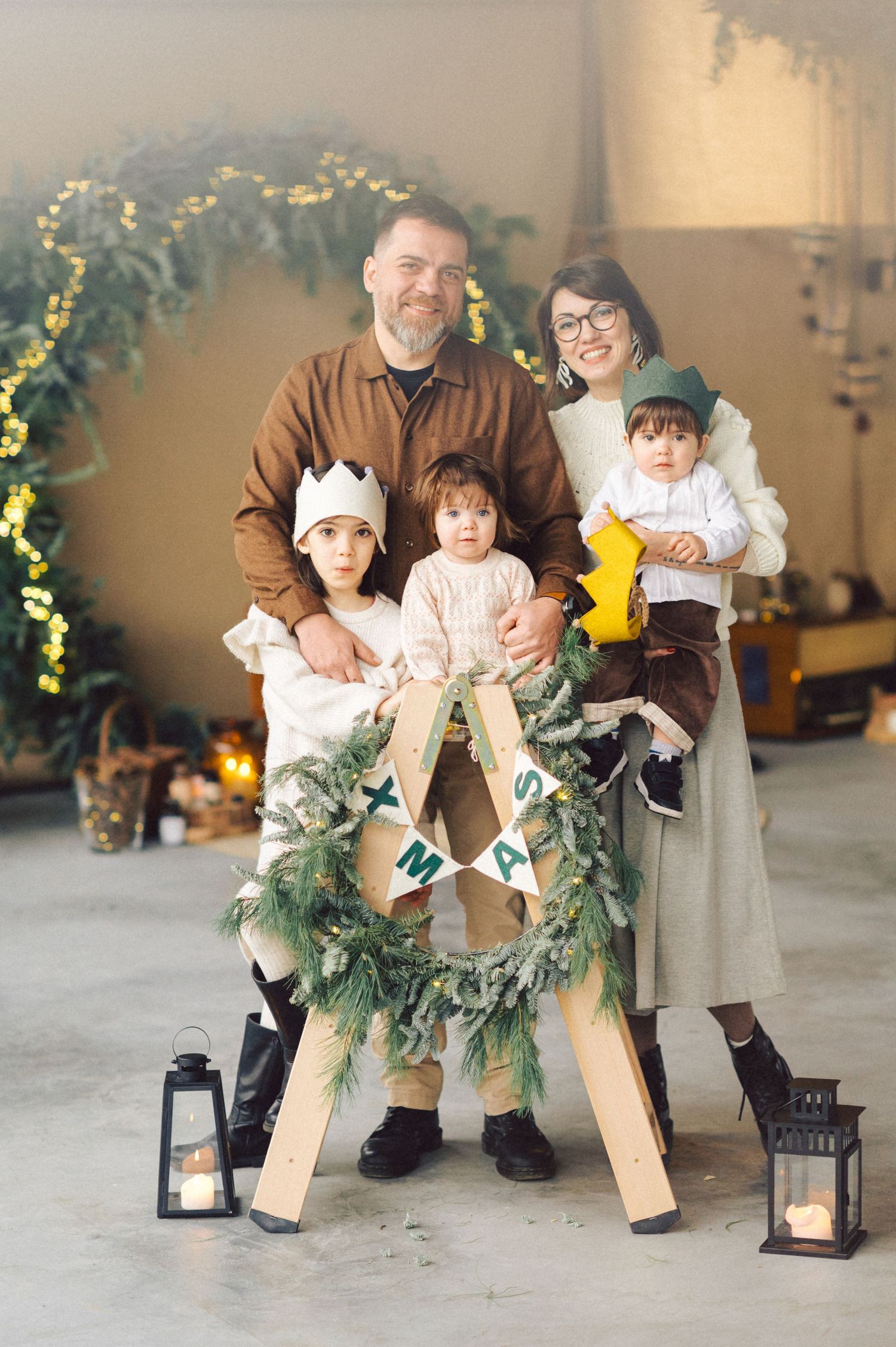 Family holiday photo session with evergreen wreath and lantern decorations in a rustic indoor setting.