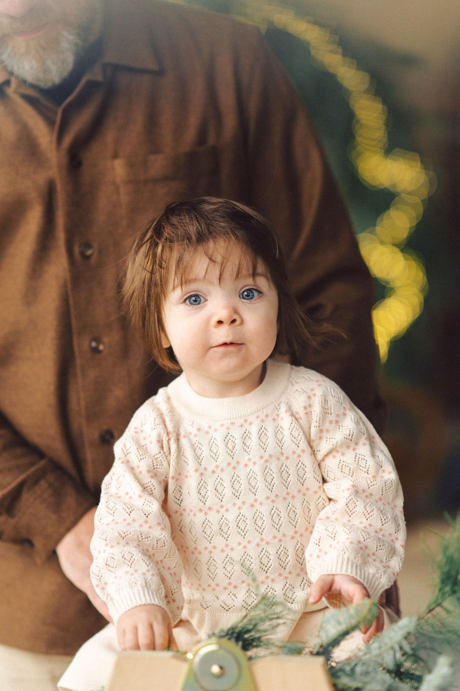A small child wearing a cream knit sweater sits beside someone in a brown shirt during a holiday photo shoot.