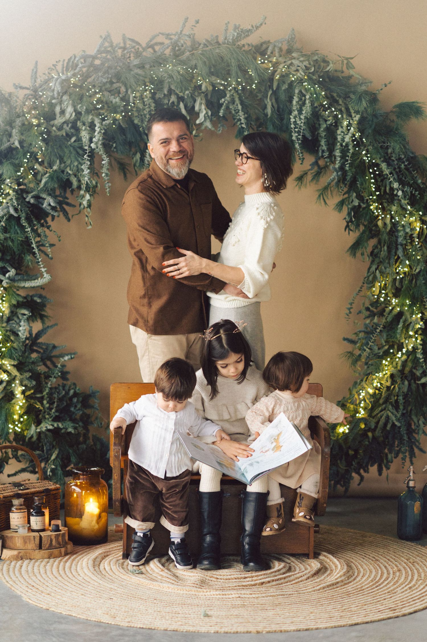 A family sits together on a round woven rug under decorative greenery garland with glowing candles in the background.