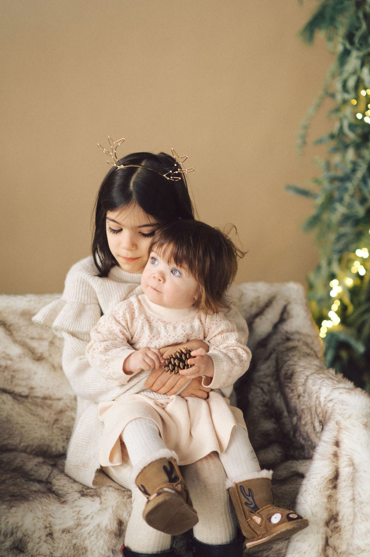 A young girl in a cream dress sits on a fur blanket examining a pinecone during a holiday photoshoot.