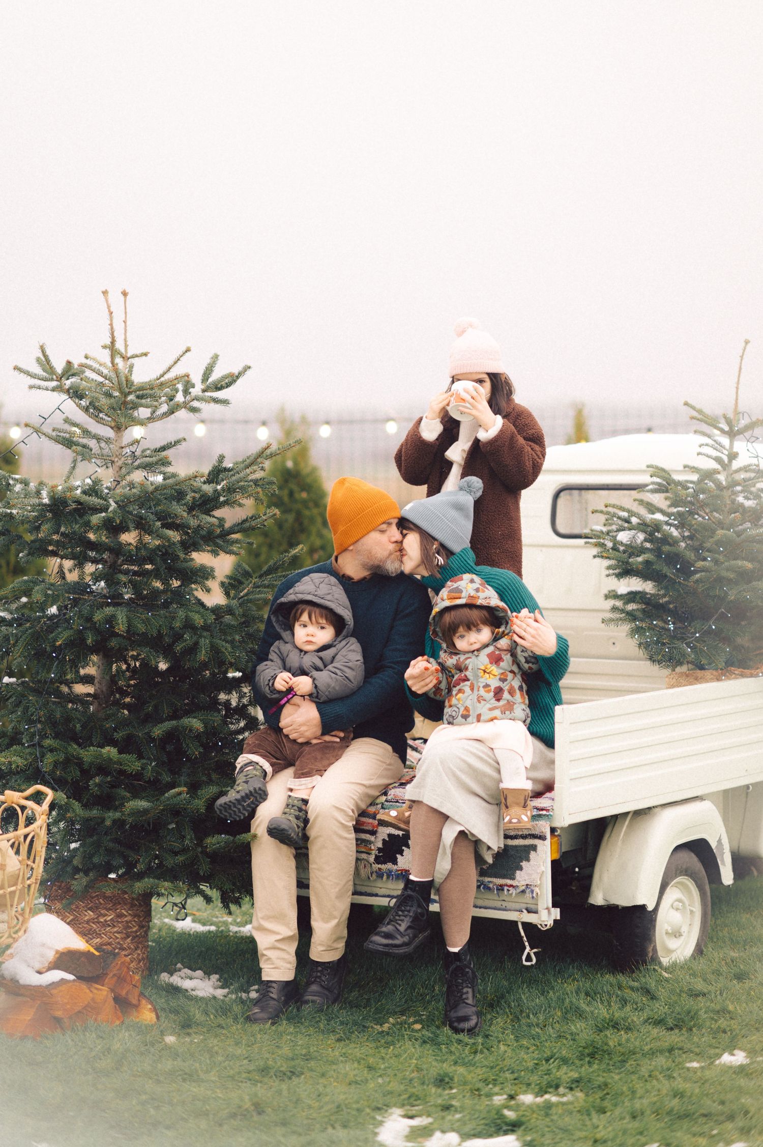 A family sits together on a white pickup truck between two evergreen Christmas trees at a holiday tree farm.