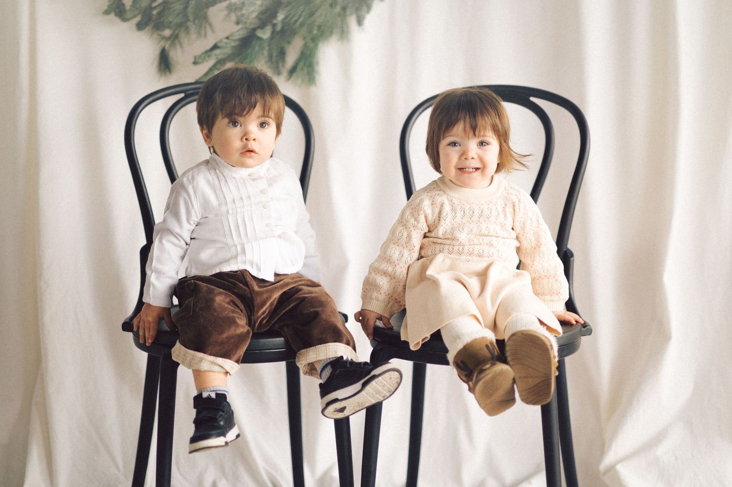 Two small children wearing light colored tops sit side by side on black chairs against a white curtain backdrop.