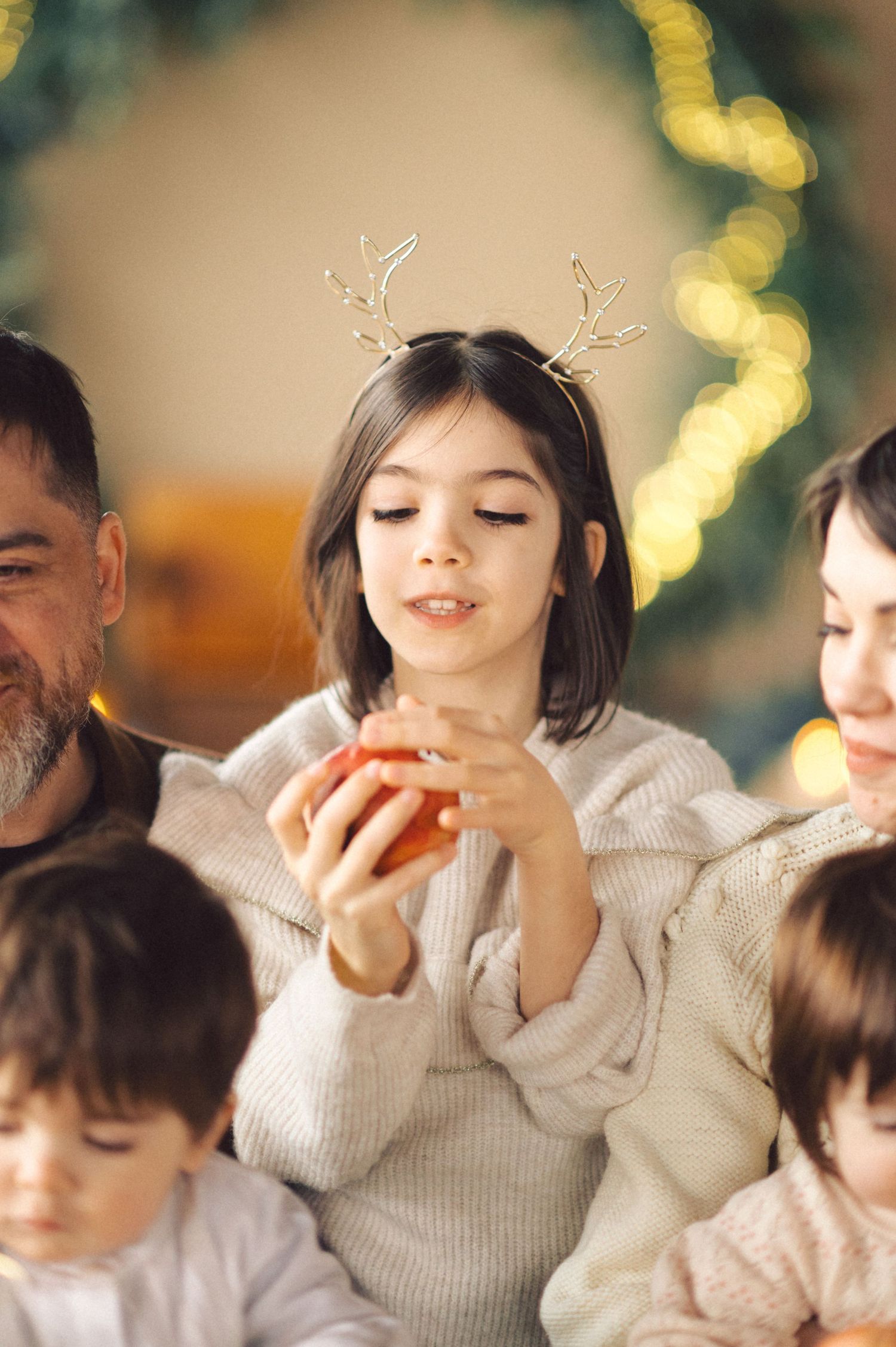 Family in cozy winter attire enjoys holiday celebrations with antler decorations and warm lighting in background.