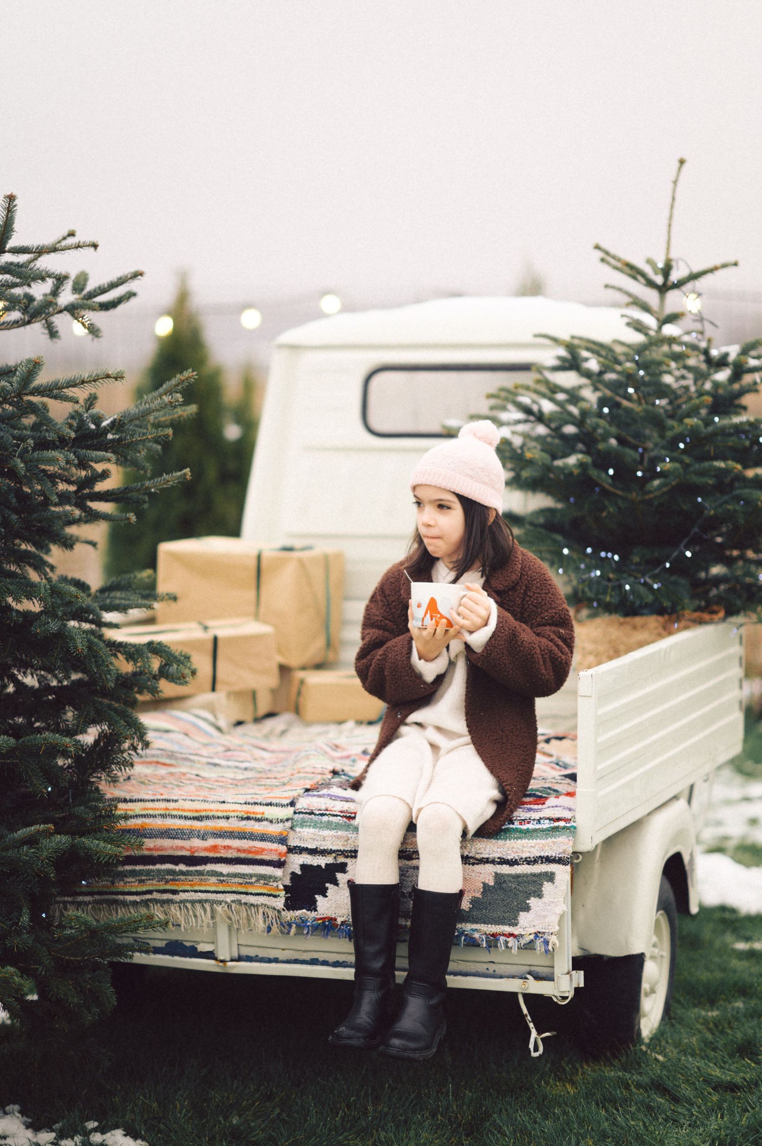 A person in winter attire enjoys a hot drink while sitting on a bench among evergreen trees in a snowy setting.