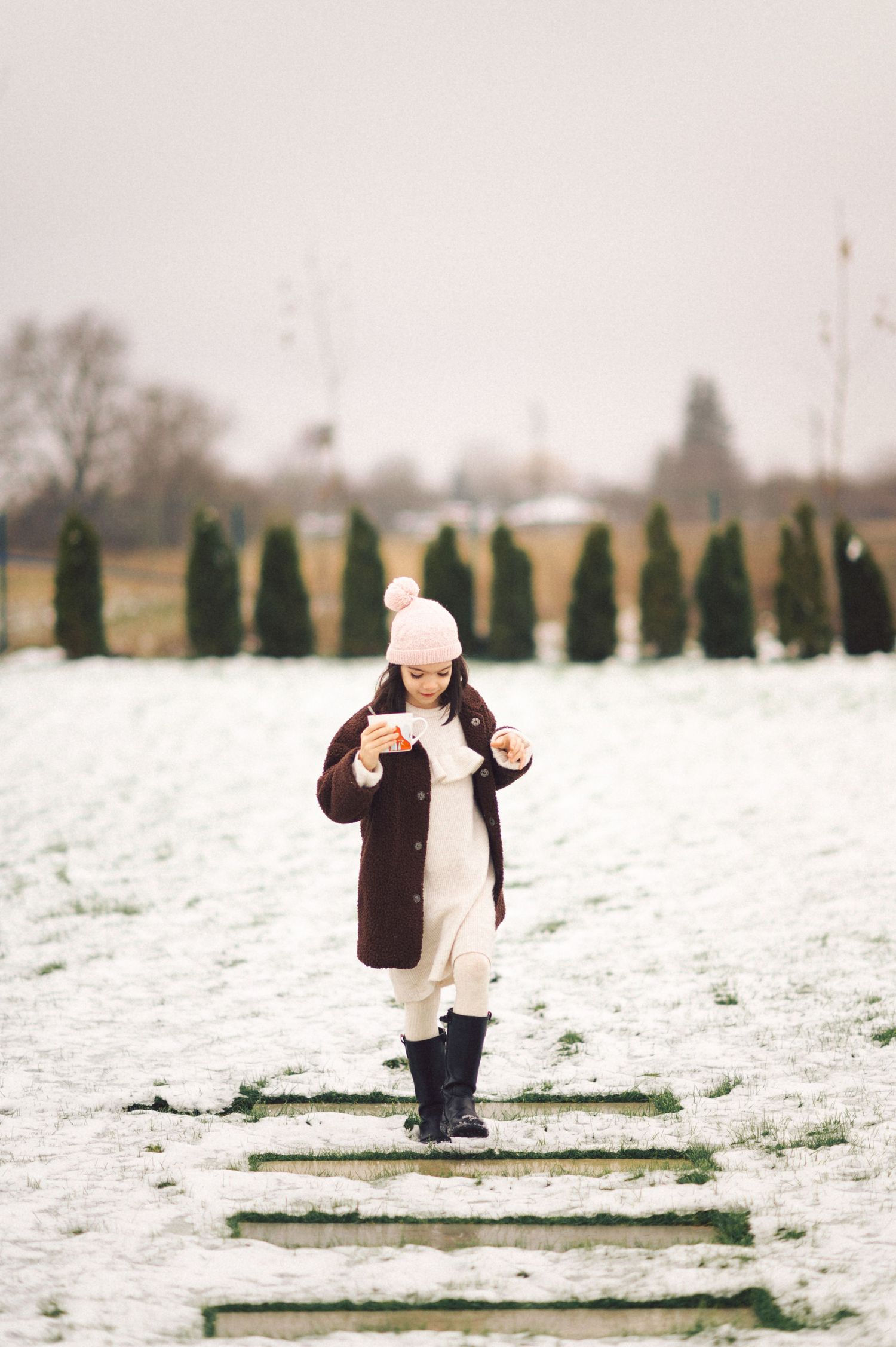 A person walks along snowy paving stones wearing a brown coat, white hat, and boots with evergreen trees in the background.