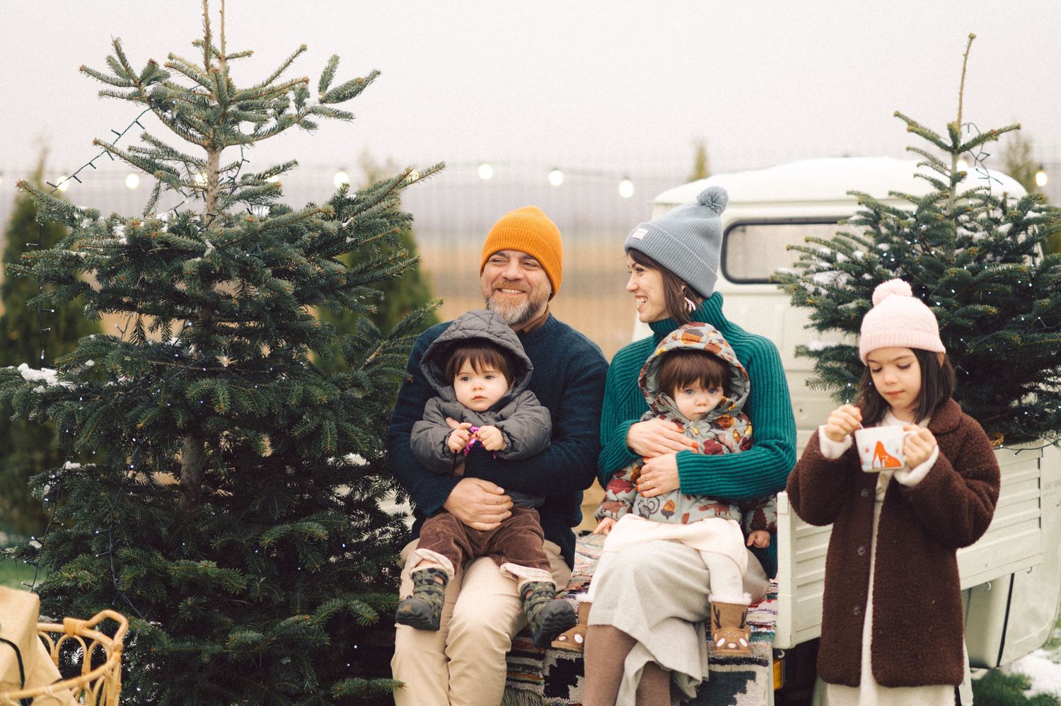 Group bundled in winter attire sits among Christmas trees at a tree farm with vintage car in background.