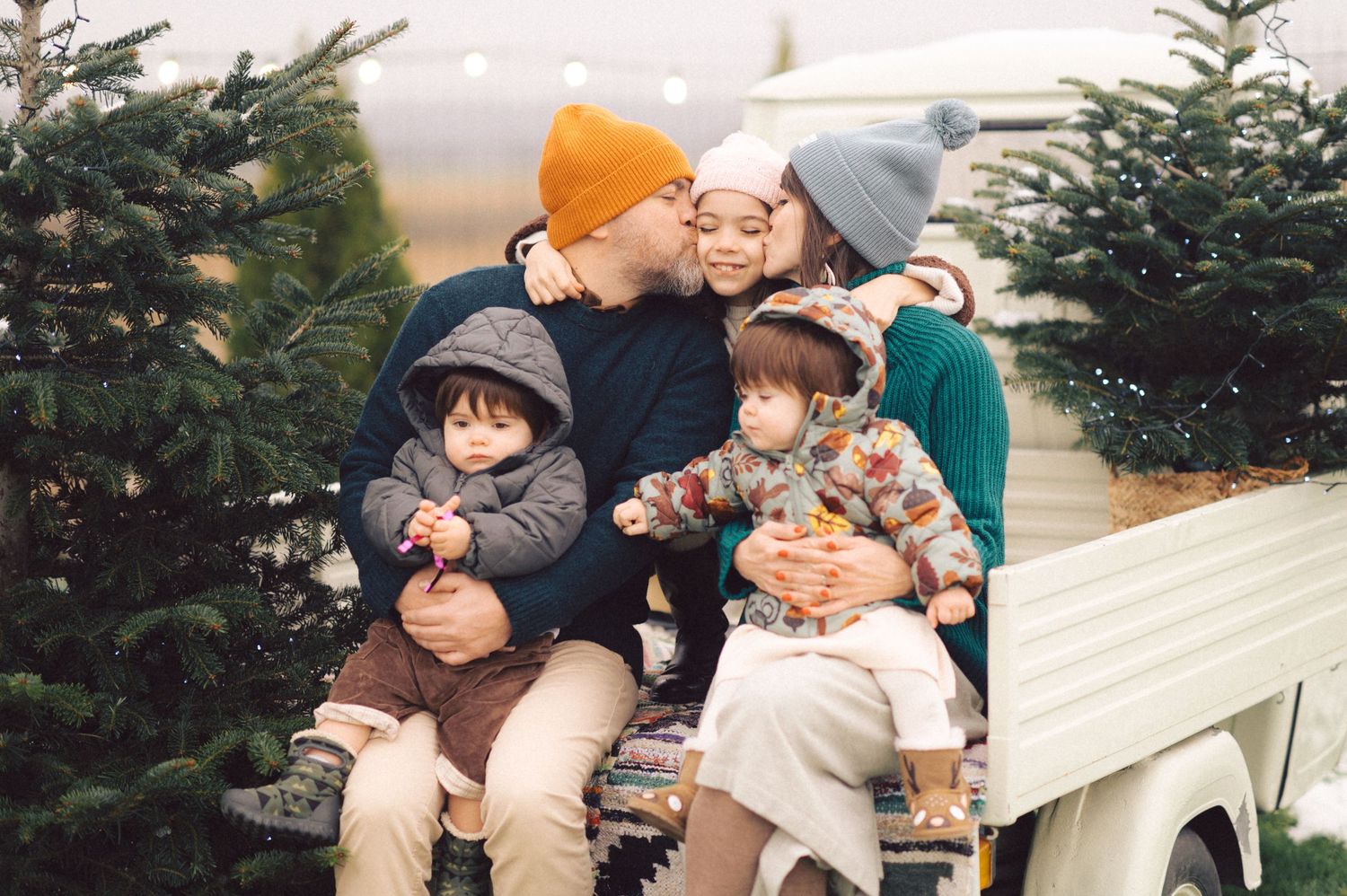 A family sits together on a bench among evergreen trees during winter, wearing cozy knit hats and warm clothing.