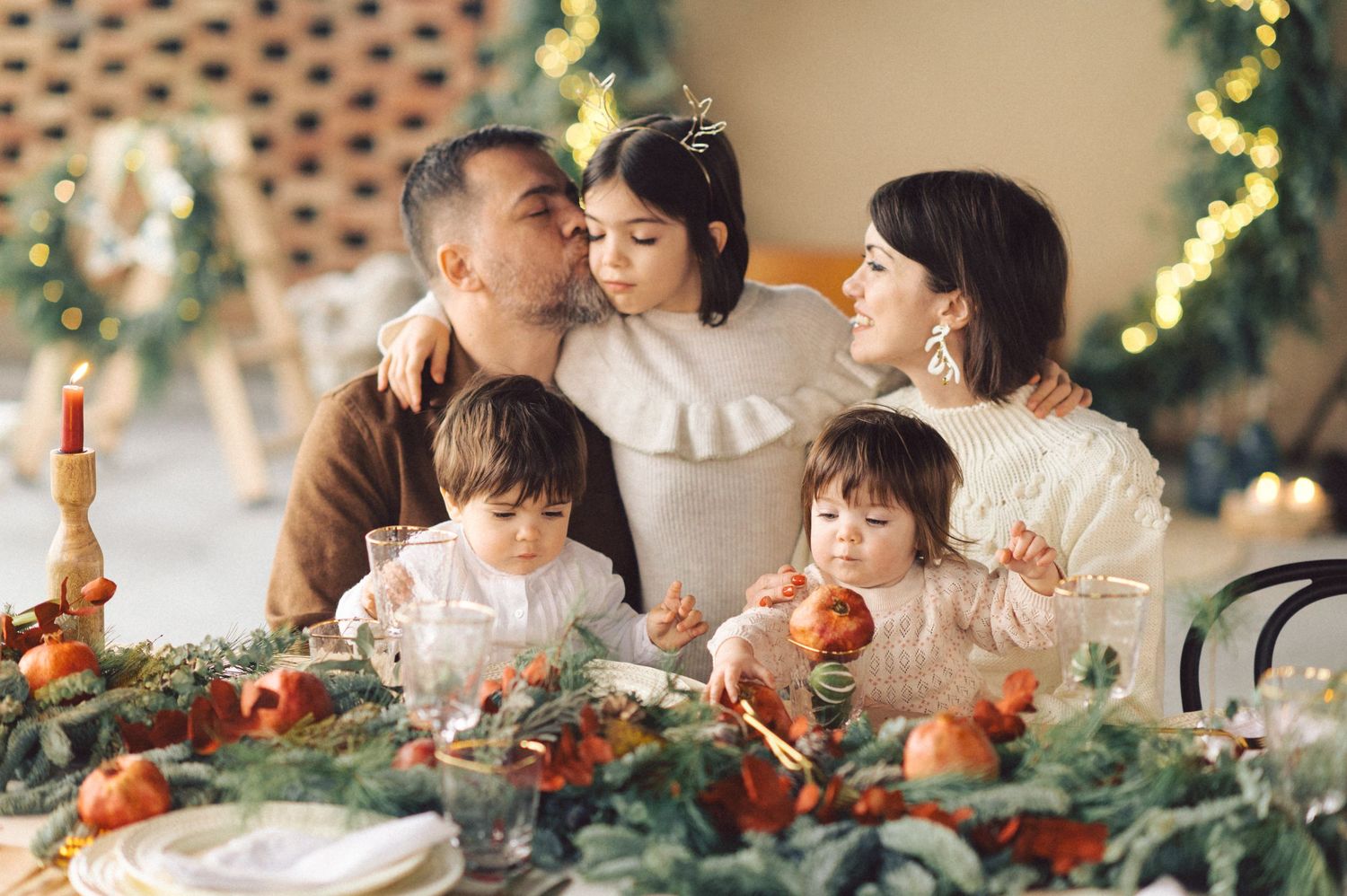 A family gathers around a festive Christmas table decorated with greenery, candles and twinkling lights in the background.