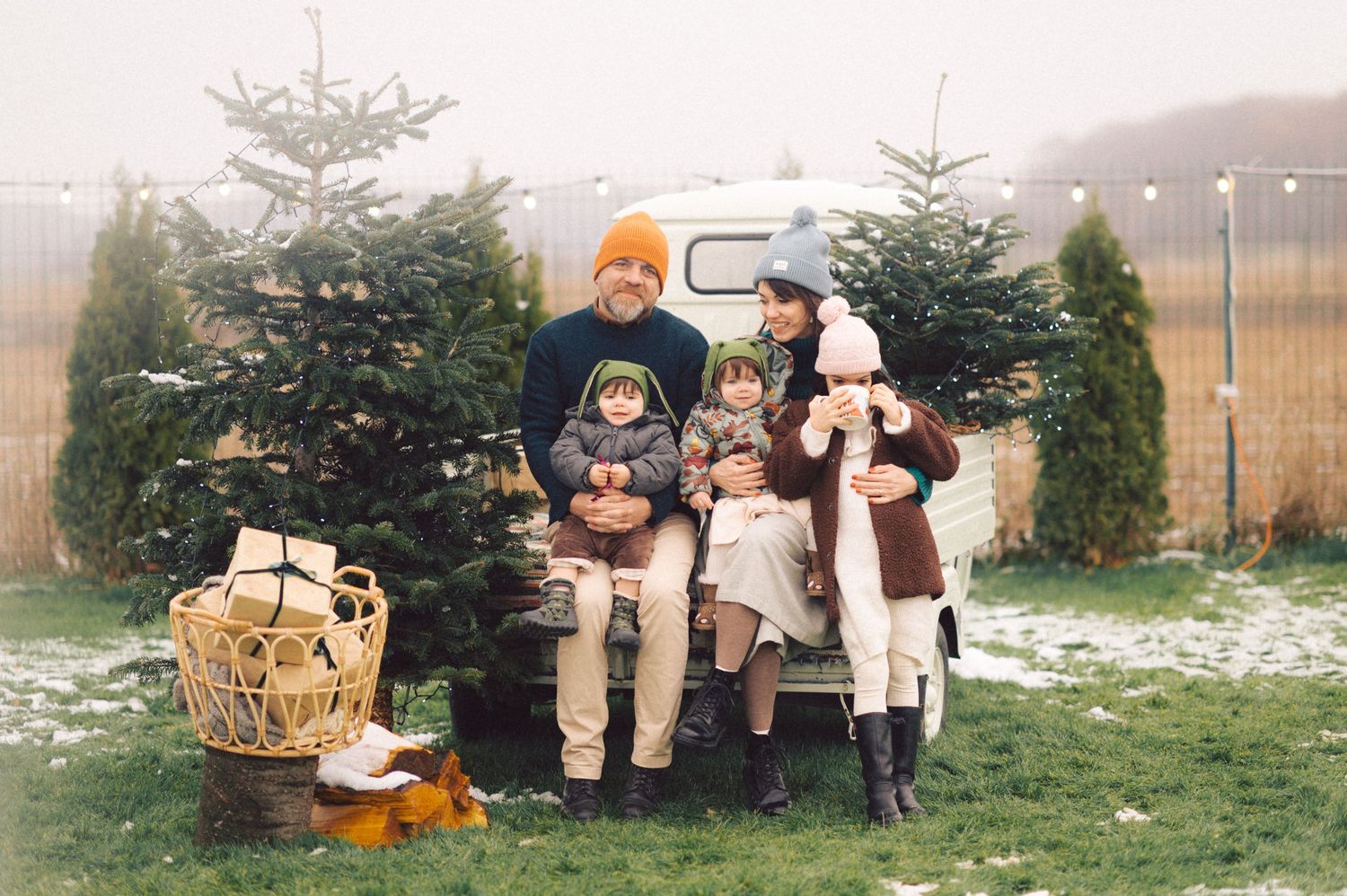 Winter family portrait with evergreen trees and rustic baskets in a misty outdoor setting.