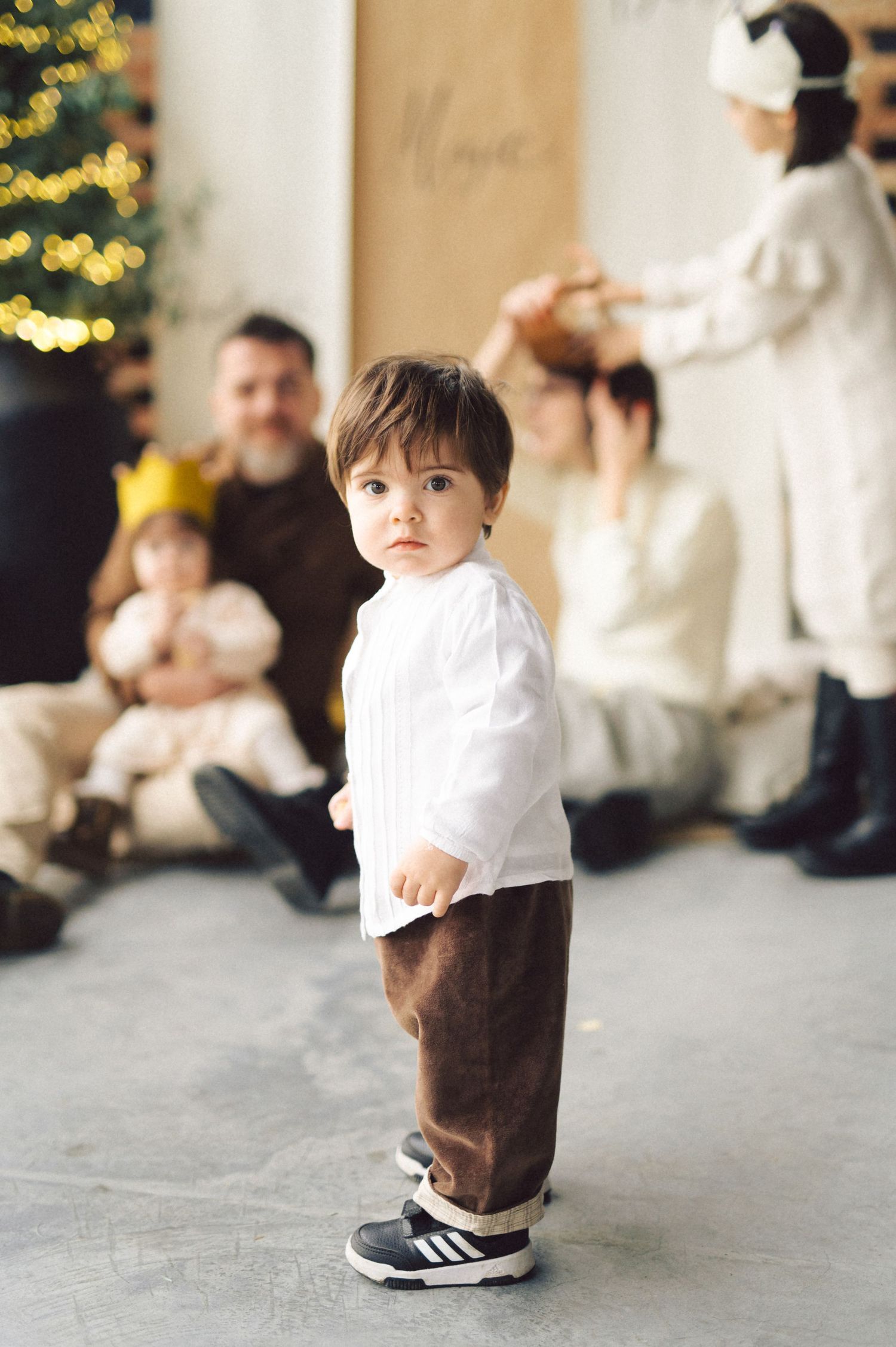 A child in a white shirt and brown pants stands in front of a Christmas tree while family gathers in the background.