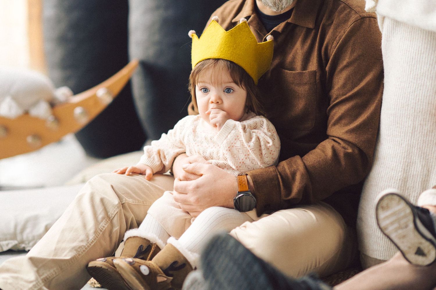 Baby wearing yellow paper crown sits with parent on white couch in casual indoor setting.
