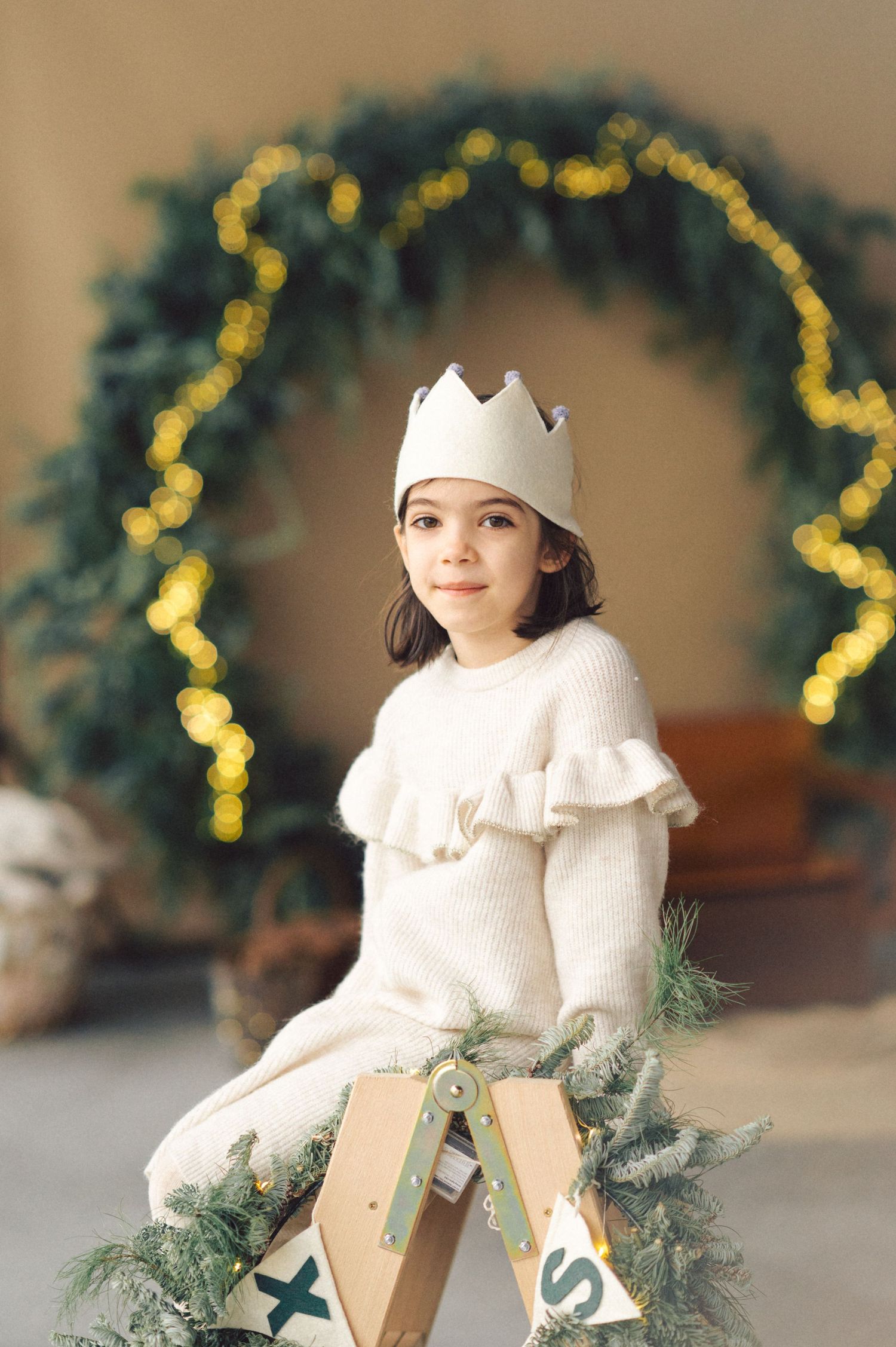 A child in a white winter sweater and paper crown sits on a wooden stool with holiday decorations and lights in the background.