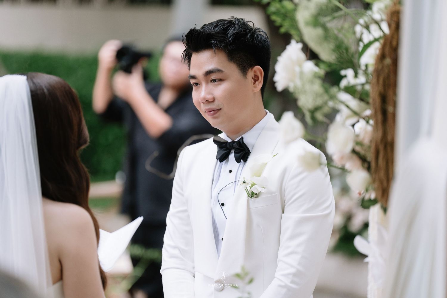 Handsome groom in white tuxedo with black bow tie smiles warmly during outdoor wedding ceremony.