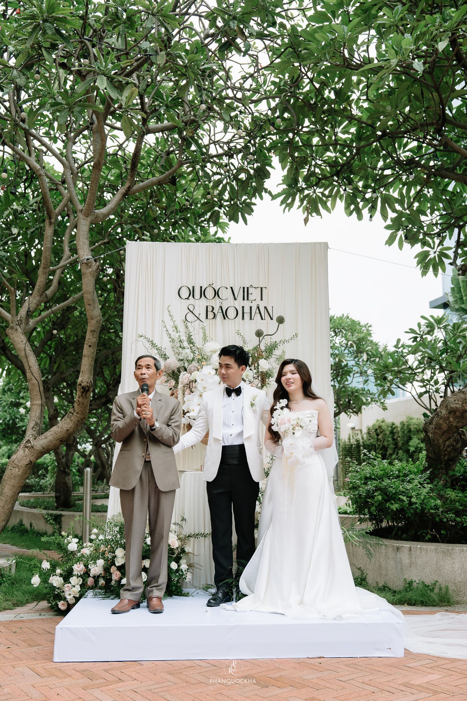 Bride and groom stand with officiant at outdoor wedding ceremony before elegant white floral backdrop reading Quoc Viet and Bao Han.