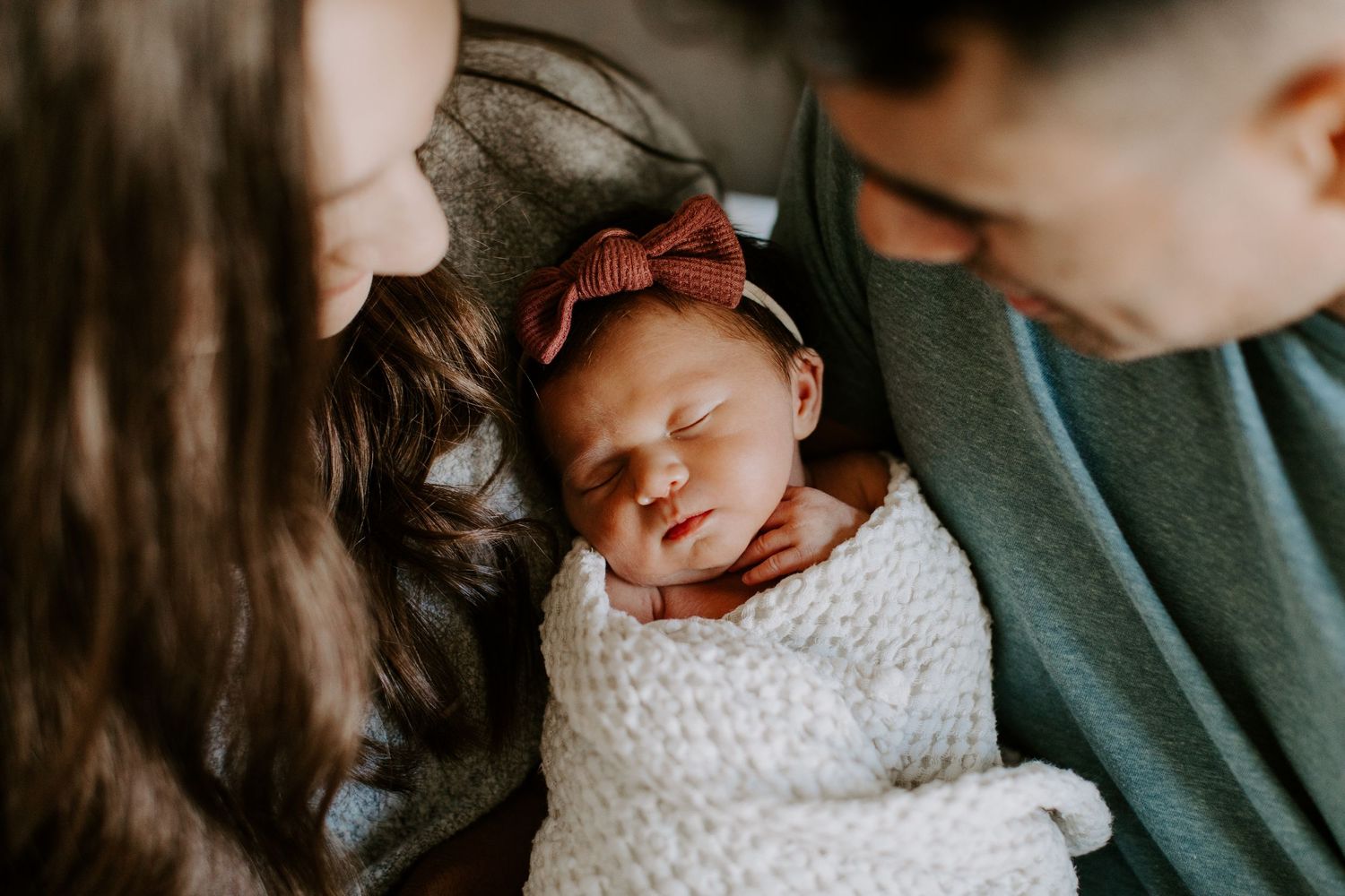 A sleeping newborn baby wearing a red bow headband is nestled between parents wearing white and green.