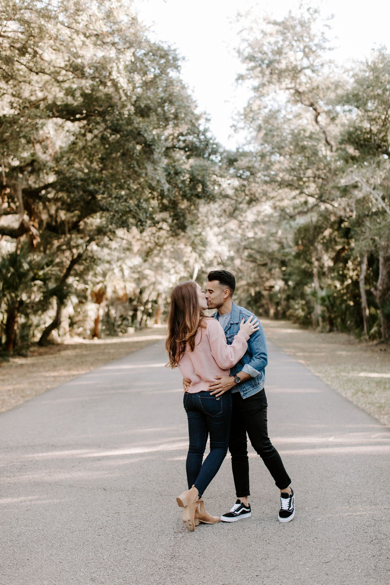 A romantic couple shares a tender moment dancing together on an empty tree-lined road at sunset.
