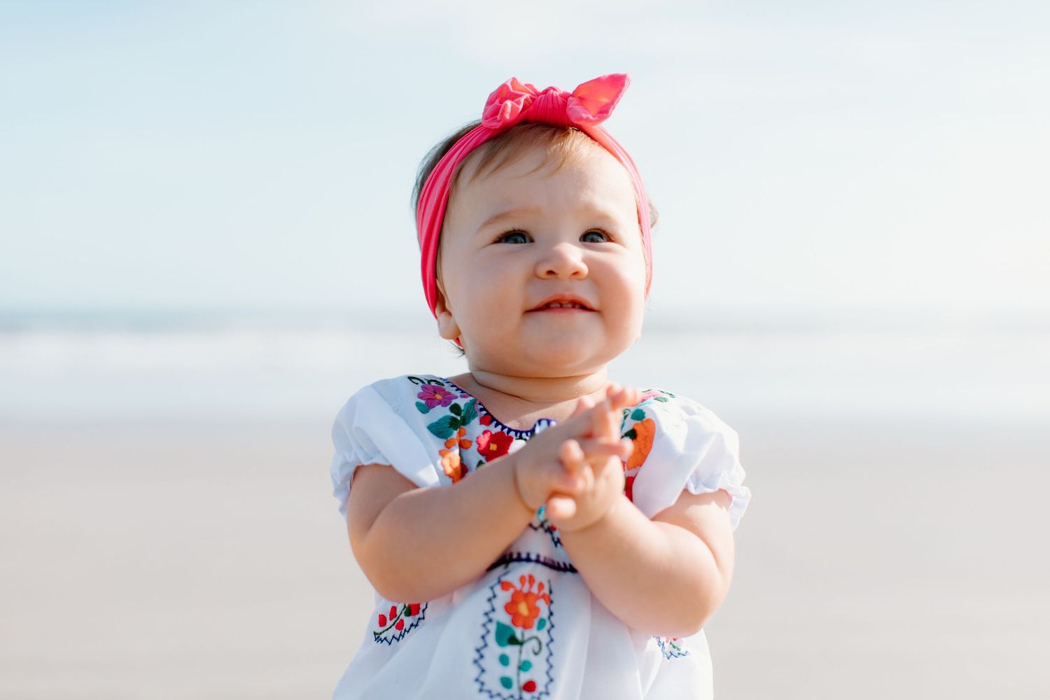 A baby wearing a red bow and floral outfit smiles while playing outdoors in bright natural light.