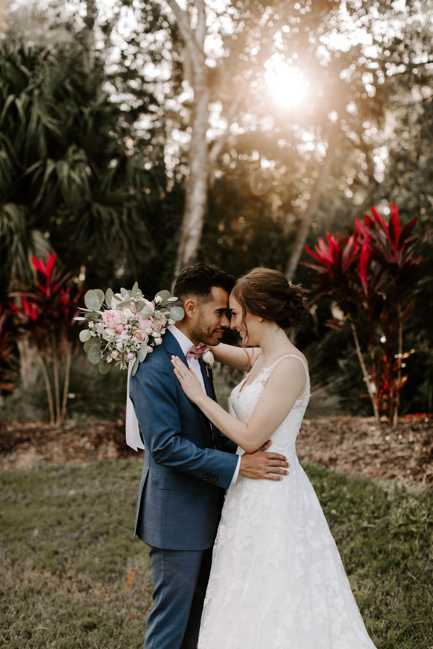 A romantic wedding portrait in a tropical garden setting with palm trees and red flowers during golden hour sunset.