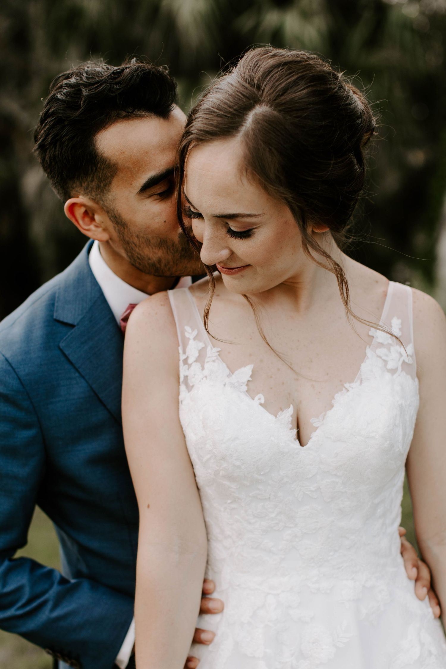 A romantic wedding portrait of a couple sharing a tender moment outdoors with the bride in a lace white dress.