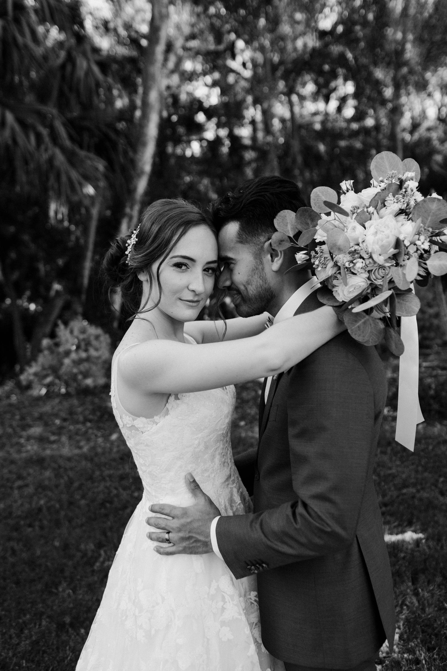 An intimate black and white wedding portrait of a couple embracing in a garden setting with a delicate floral bouquet.
