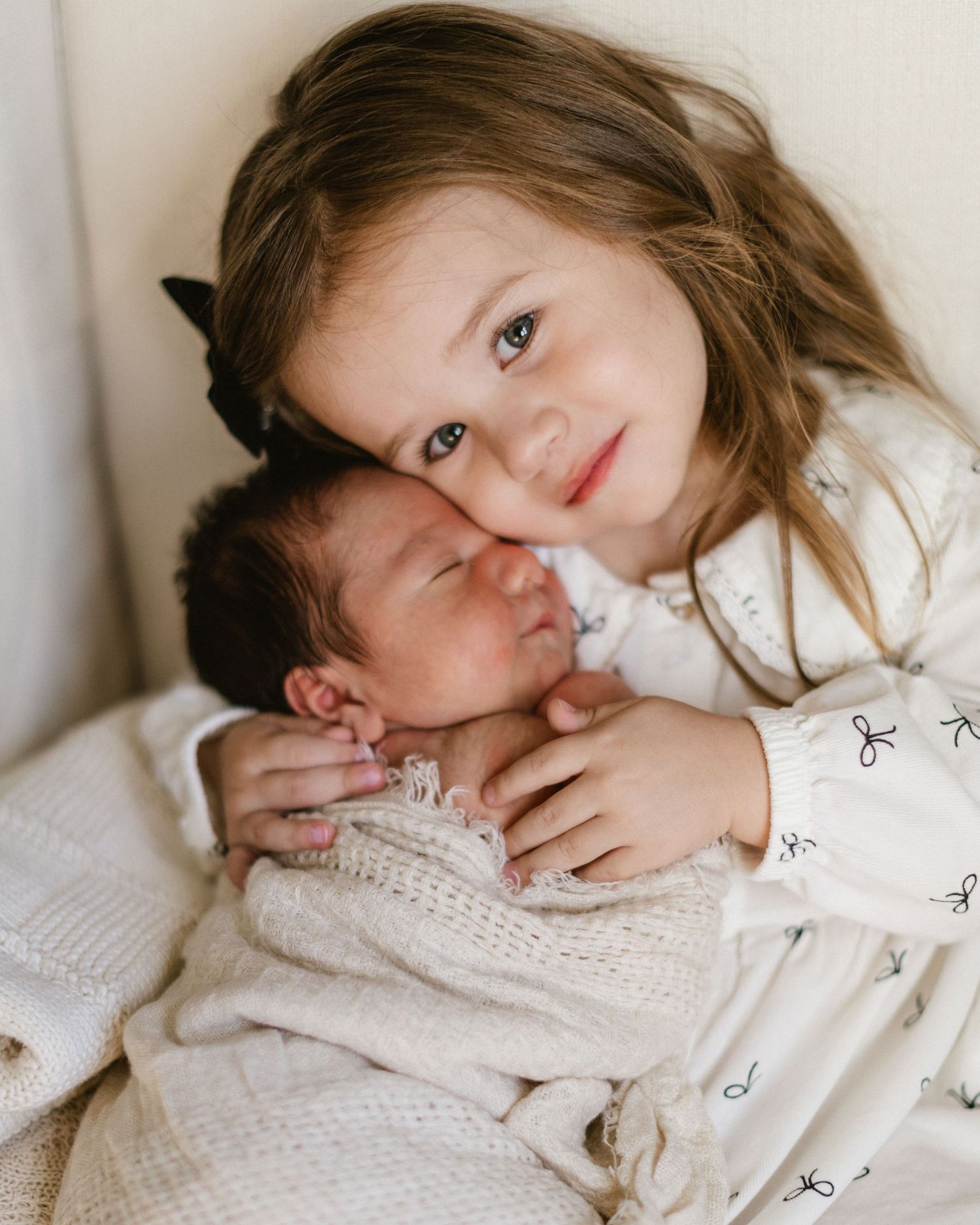 A young child embraces a sleeping newborn baby wrapped in knit blanket while laying on soft white bedding.