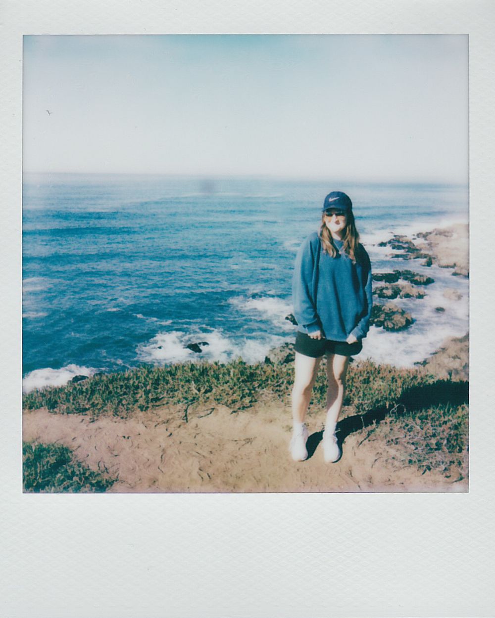 A person in a blue jacket sits on a coastal cliff overlooking the rugged Pacific Ocean shoreline.