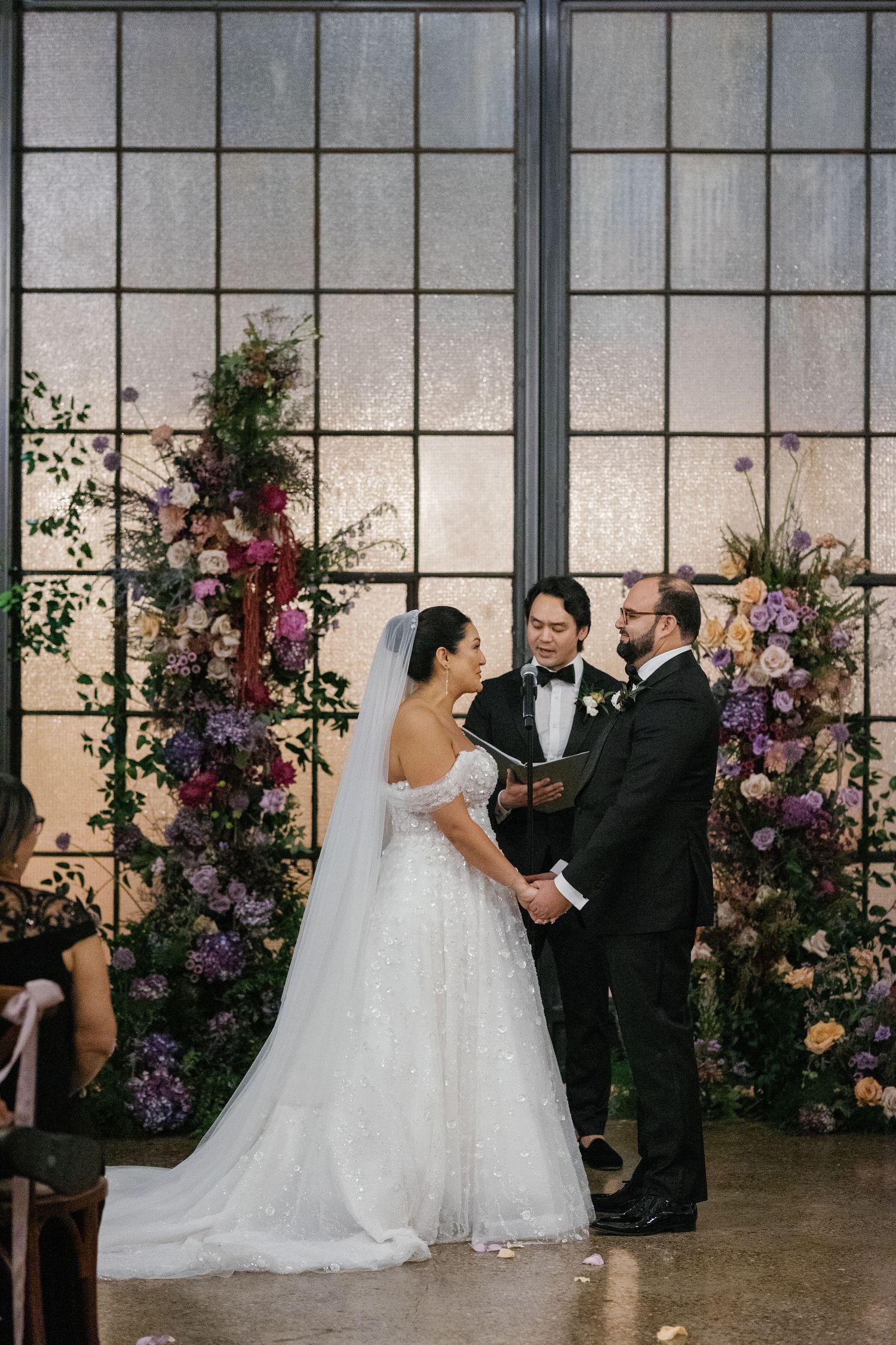 Beautiful wedding ceremony with floral arrangements and industrial windows at an indoor venue with romantic lighting.