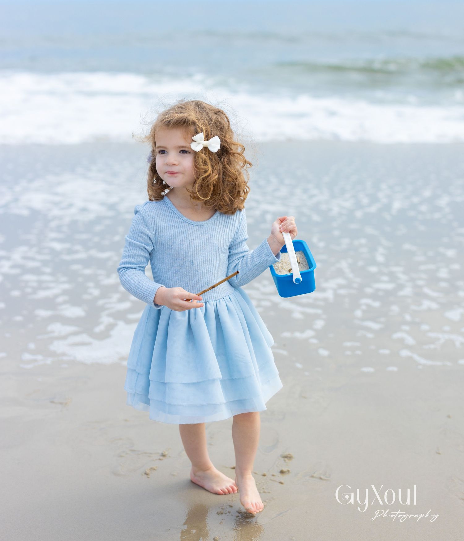 A young child wearing a light blue dress stands barefoot on a beach with ocean waves in the background.