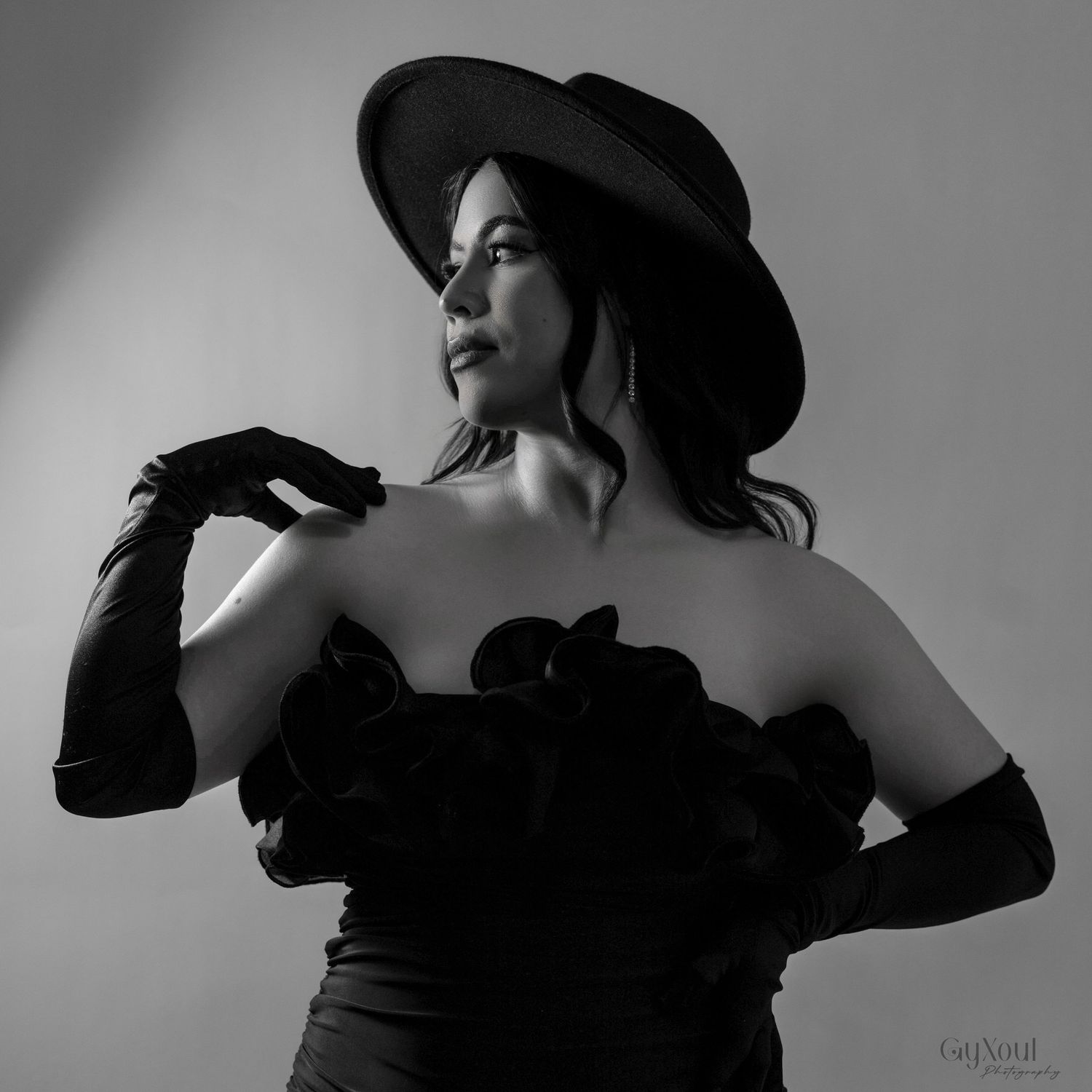 Elegant black and white portrait of a figure in hat and gloves posing dramatically against gray background.