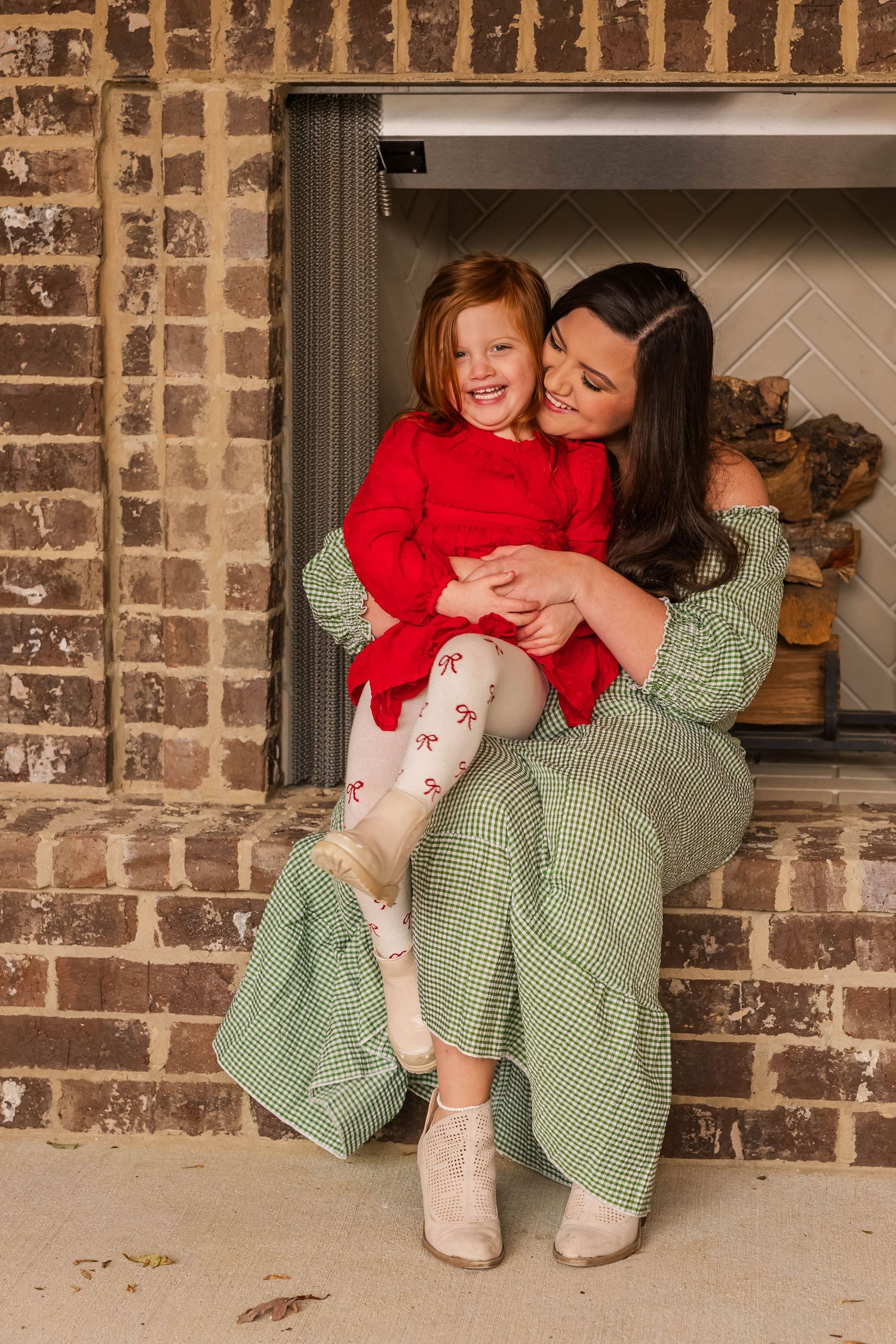 A cozy indoor portrait by a brick fireplace featuring a green gingham dress and festive red sweater.