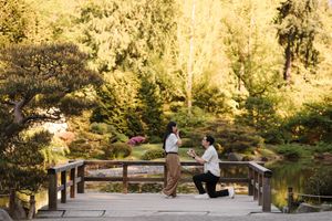 Person proposing on one knee to partner on a wooden bridge in a lush Japanese garden with golden autumn trees.