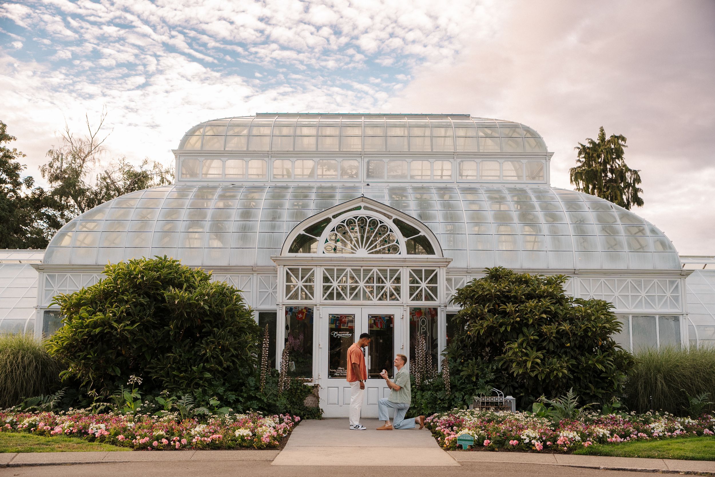 Couple standing before a grand Victorian glass greenhouse with lush gardens and a cloudy sky backdrop.