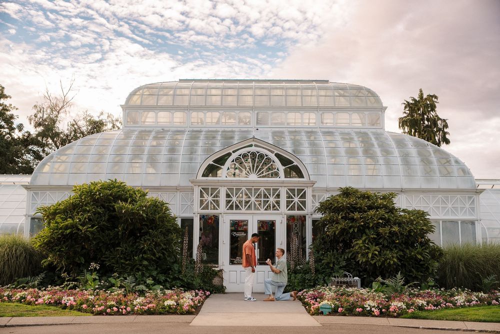 Couple standing before a grand Victorian glass greenhouse with lush gardens and a cloudy sky backdrop.
