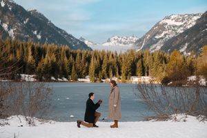 Man proposes on one knee to woman by snowy mountain lake surrounded by pine trees and snow-capped peaks in winter.