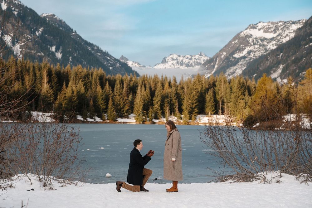 Man proposes on one knee to woman by snowy mountain lake surrounded by pine trees and snow-capped peaks in winter.