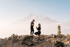 Couple celebrating a mountain top proposal with snow-capped peak backdrop on rocky summit.