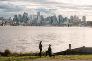 A couple shares a marriage proposal by the waterfront with a stunning city skyline backdrop on a cloudy day.