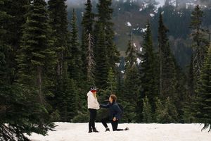 Person proposing on one knee in snowy mountain landscape surrounded by tall evergreen trees.