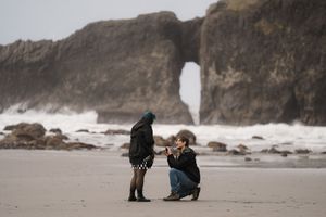 People walk along a misty beach with rugged cliffs and crashing waves in the background.