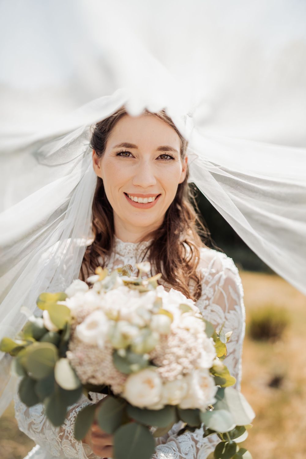 Joyful bride wearing white lace dress and veil holds beautiful white rose and eucalyptus bouquet while smiling outdoors.