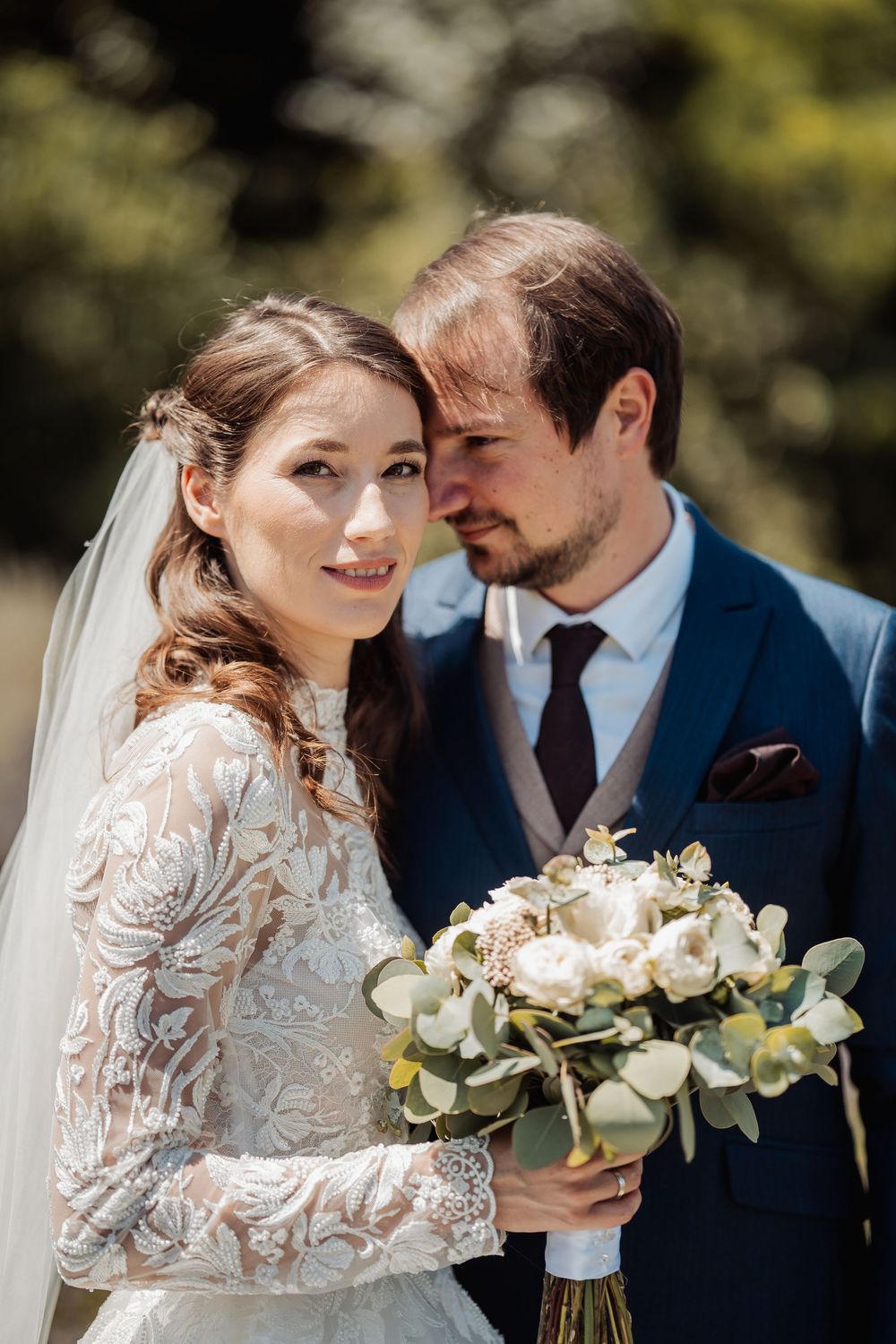 Bride in lace long sleeve dress holds white bouquet next to groom in navy suit on sunny outdoor wedding day.