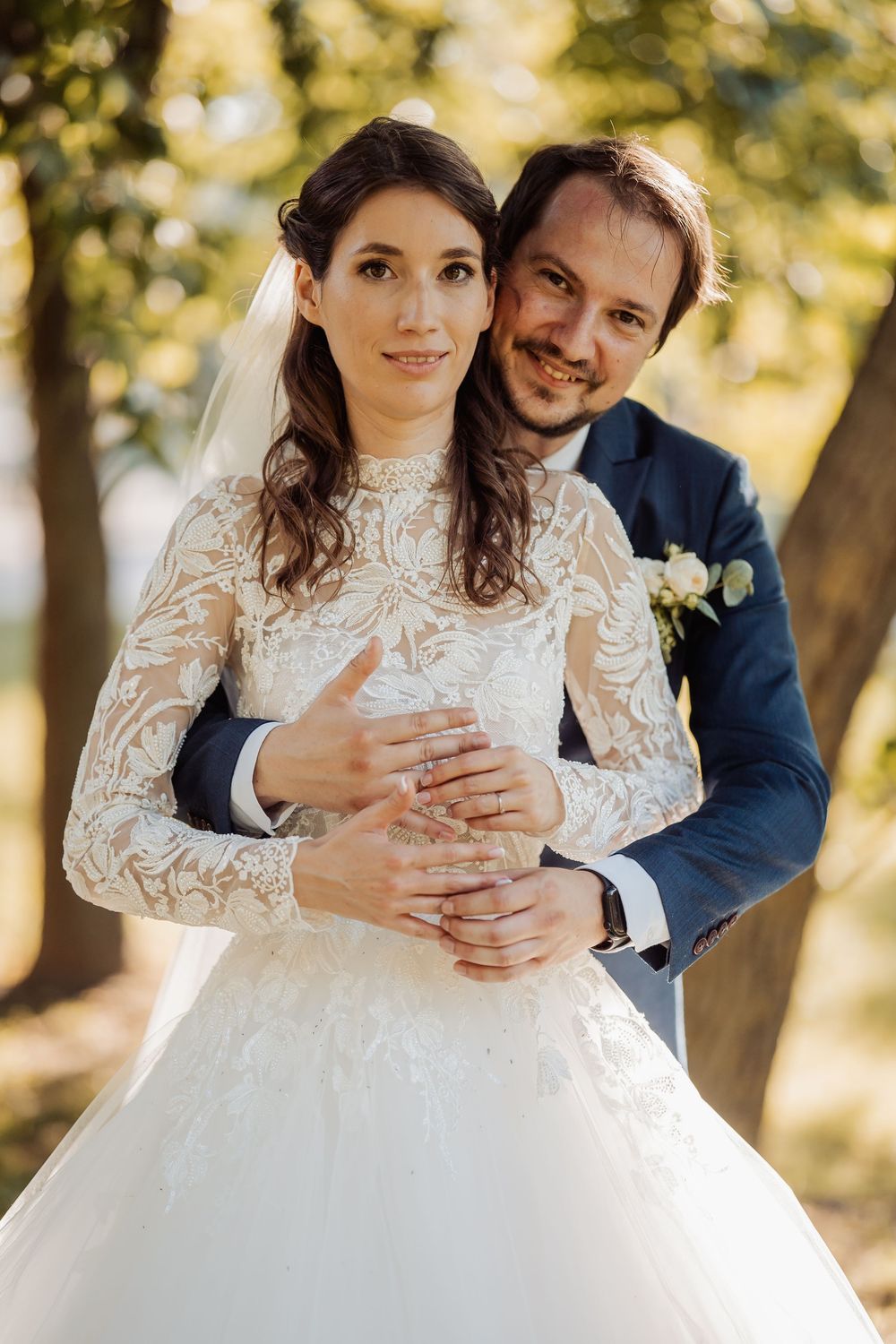 A wedding couple poses outdoors in autumn sunlight, bride wearing long sleeve lace gown and groom in navy suit.