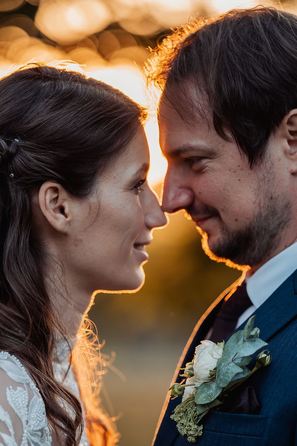 A romantic couple in wedding attire share an intimate moment at sunset with golden backlighting creating a warm glow.