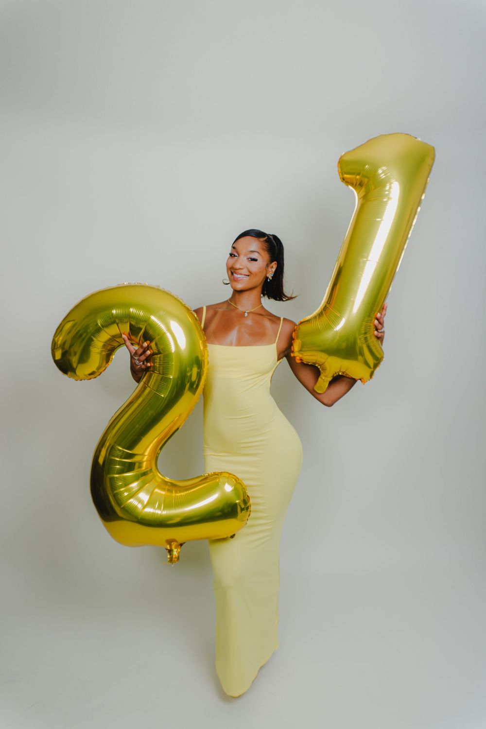 Model in yellow dress poses playfully with gold number balloons against white background.