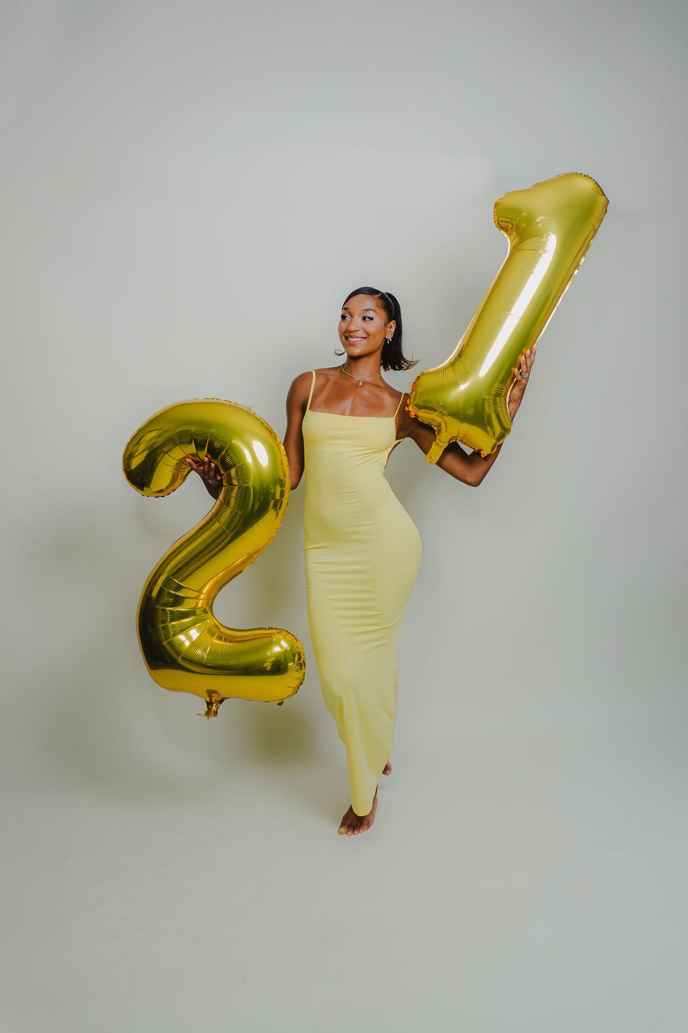Joyful poses with gold number balloons and fitted yellow dress in studio setting.