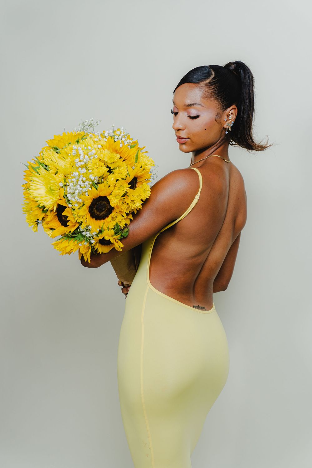 Model in yellow dress poses elegantly with vibrant sunflower bouquet against light gray backdrop.