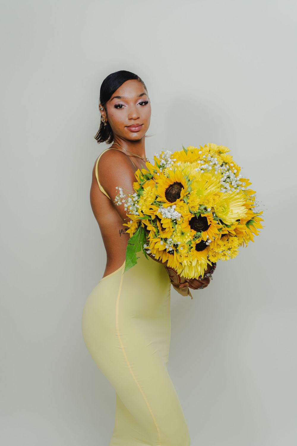 Fashion portrait with sunflowers showing model in fitted yellow dress against neutral background.