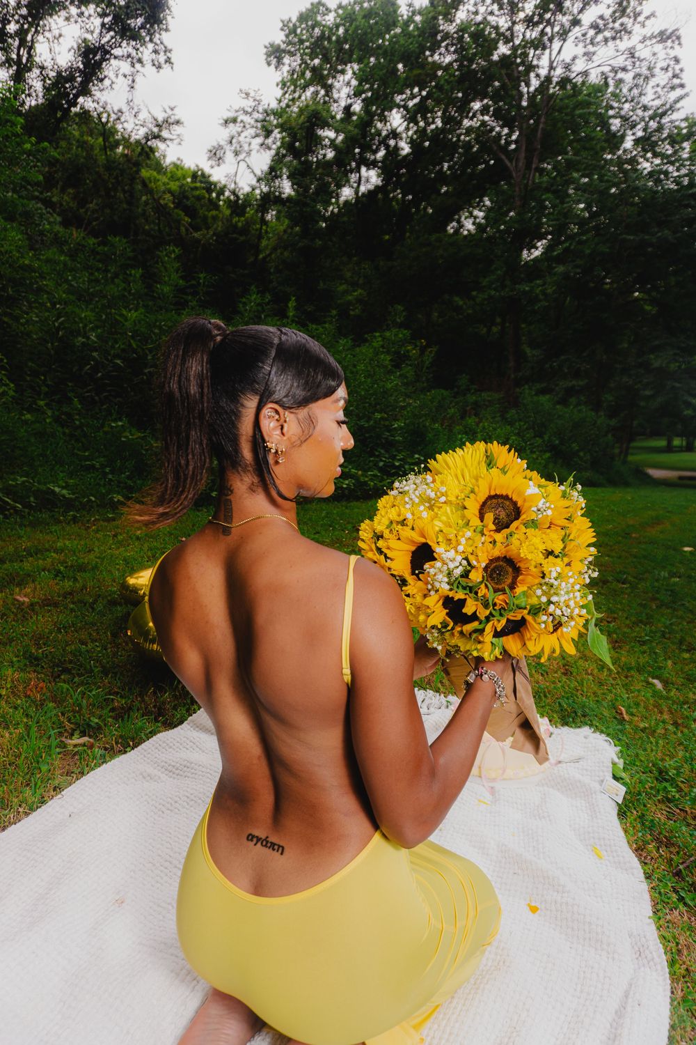 Backlit outdoor portrait with sunflower bouquet and yellow dress against forest background.