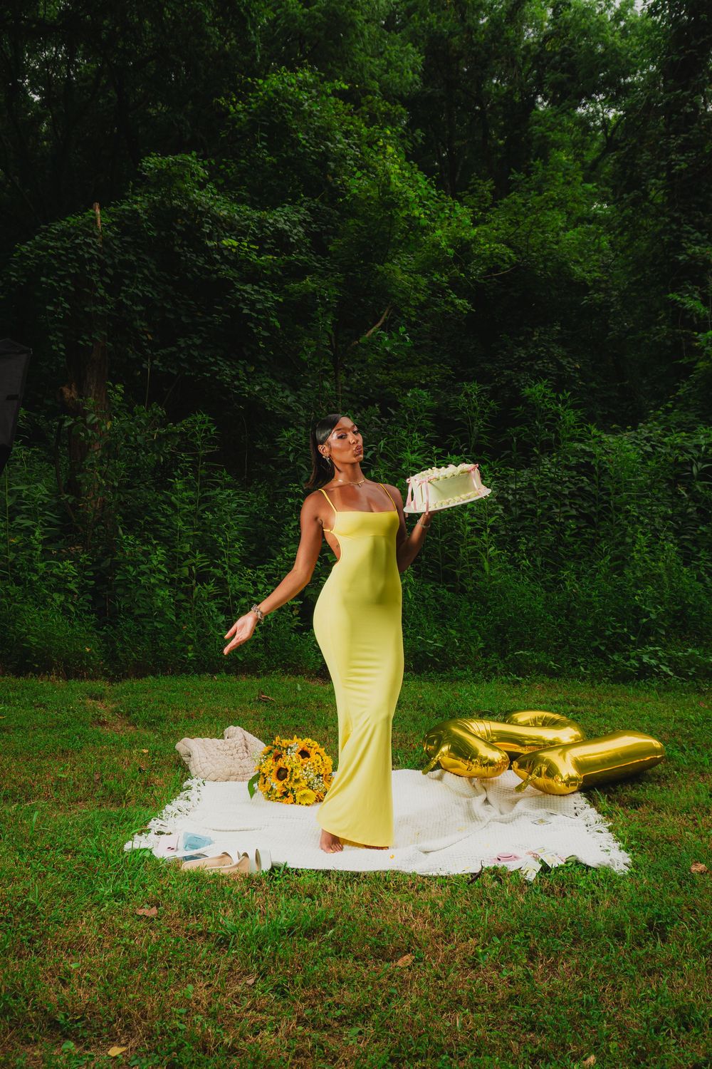 Fashion model in yellow dress poses gracefully on white blanket in grassy outdoor setting.