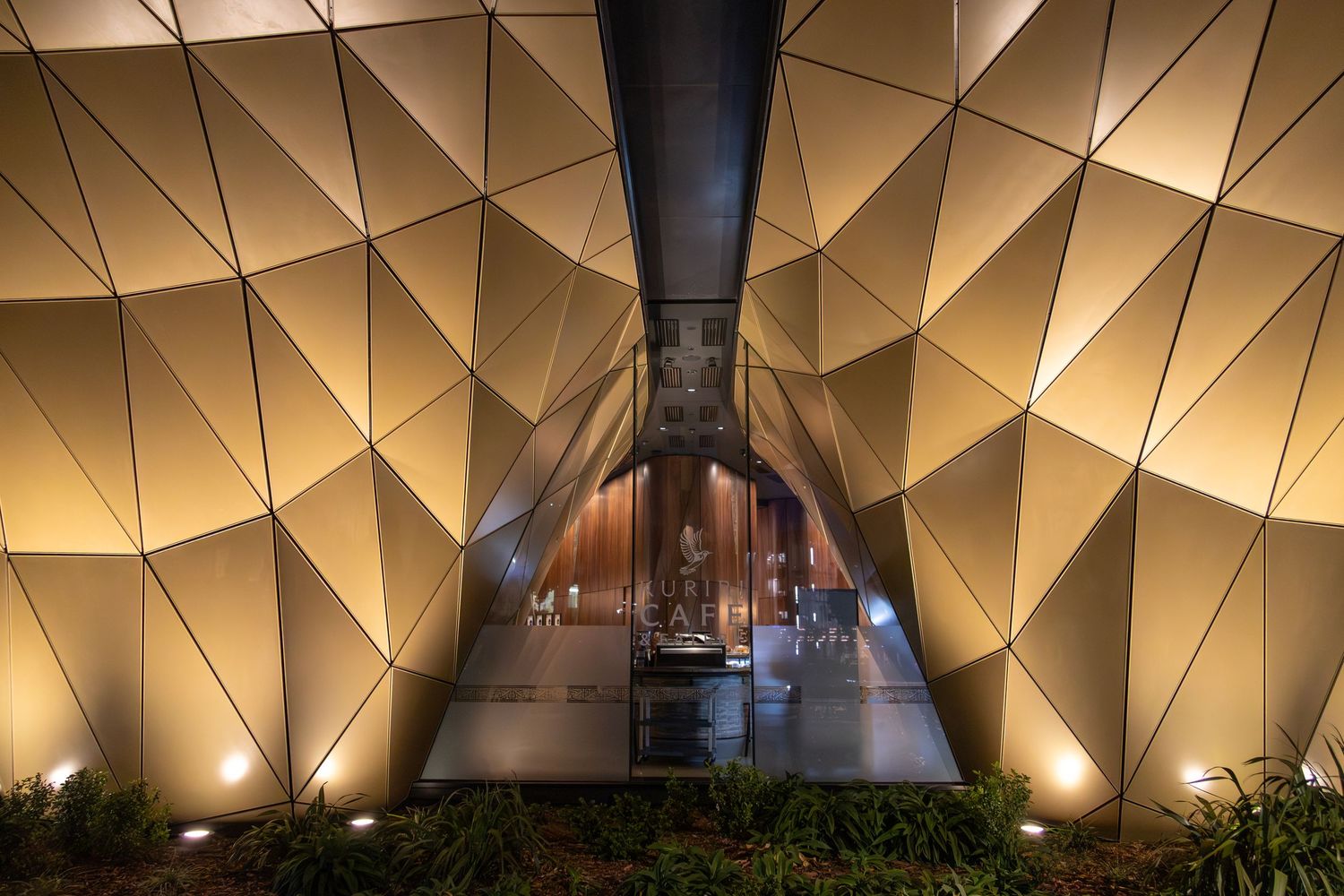 Gold-toned geometric facade forms a bold angular entrance with soft ground lighting and mirrored panels.